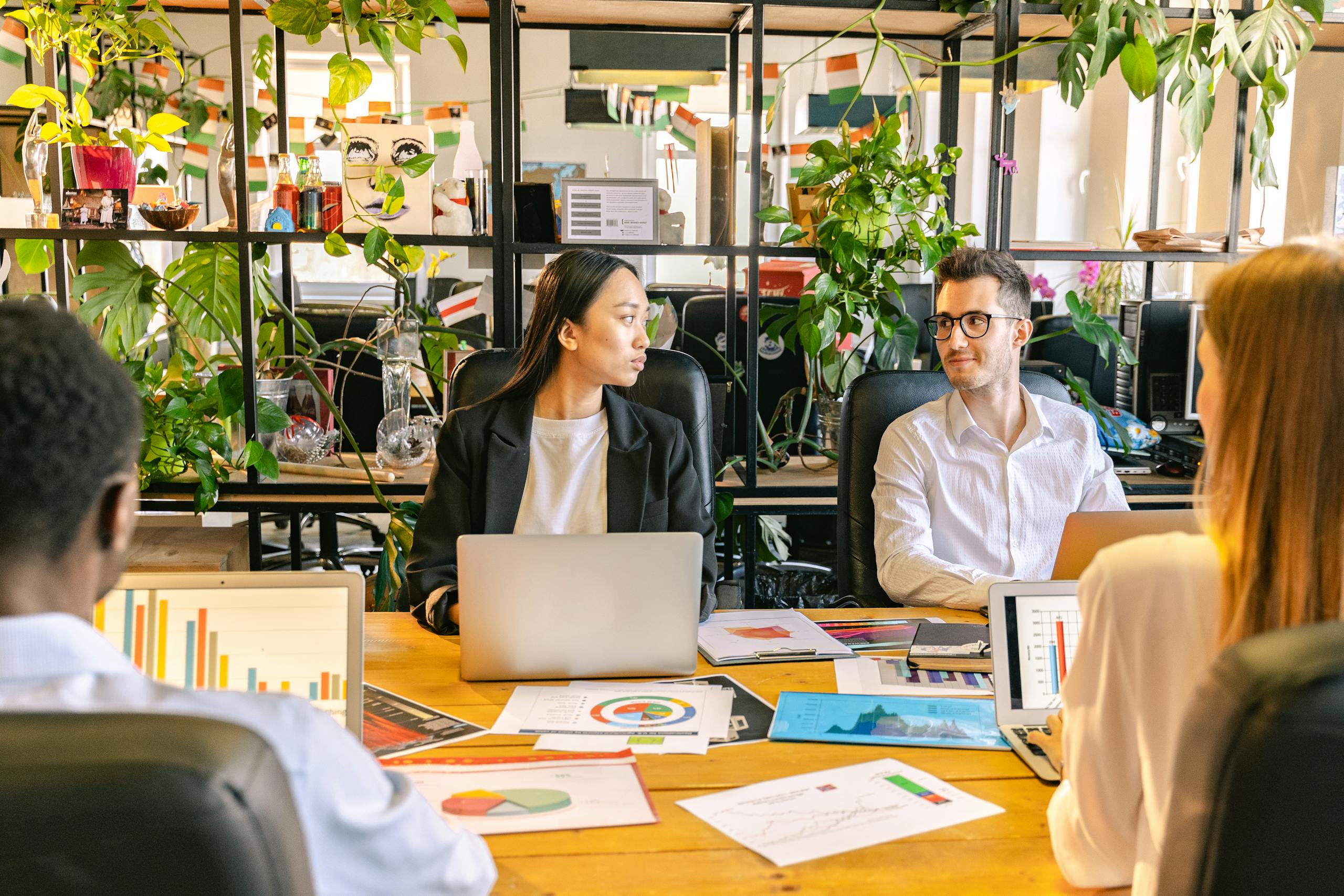 A diverse team of professionals discussing projects in a plant-filled office setting.