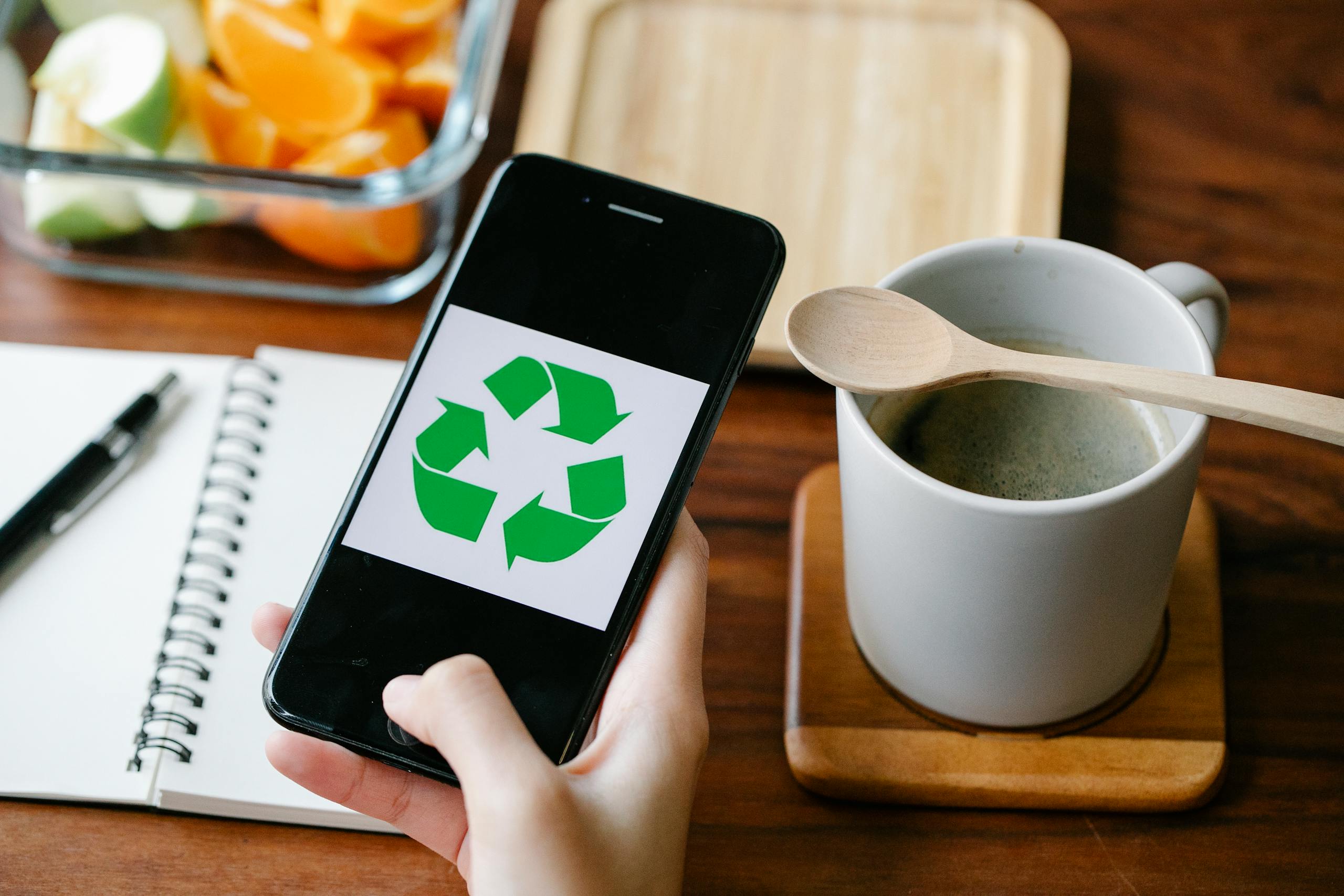 Hand holding smartphone with recycling icon beside coffee and fruit on wooden table.
