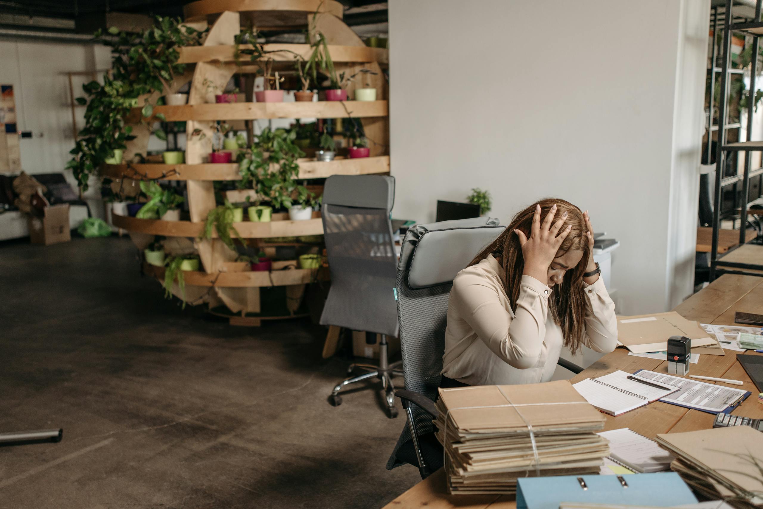 A stressed woman sits overwhelmed at her desk, surrounded by paperwork in a modern office setting.